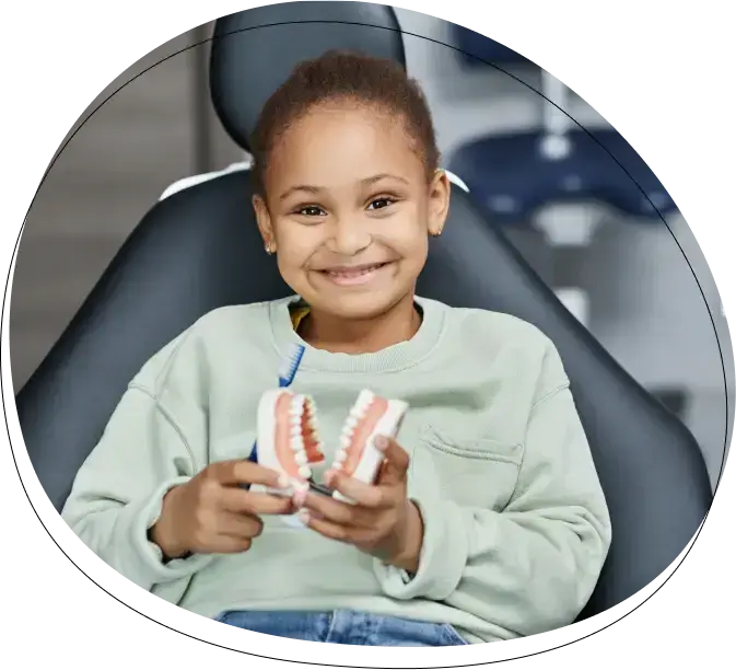 Young child smiling in dental chair holding teeth model during visit.